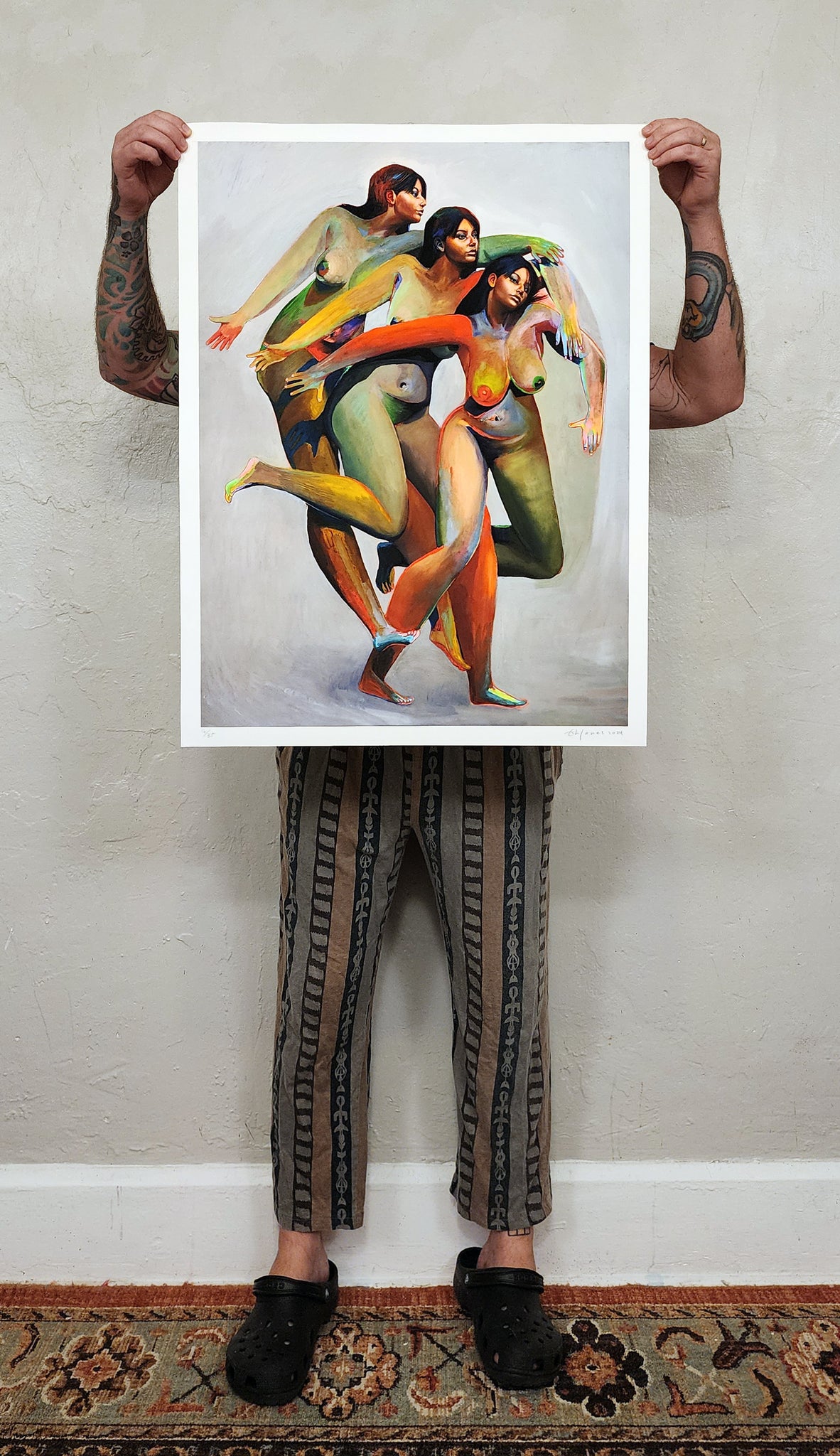 Erik Jones print of three nude figures with the same face and colorful bodies - print being held up by artist (man hidden behind print)