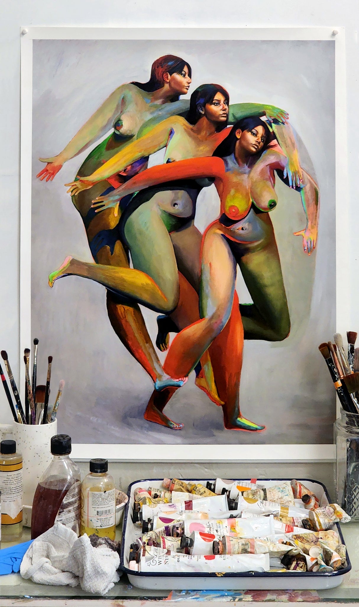 Erik Jones print of three nude figures with the same face and colorful bodies  - print hanging on wall with art supplies in foreground