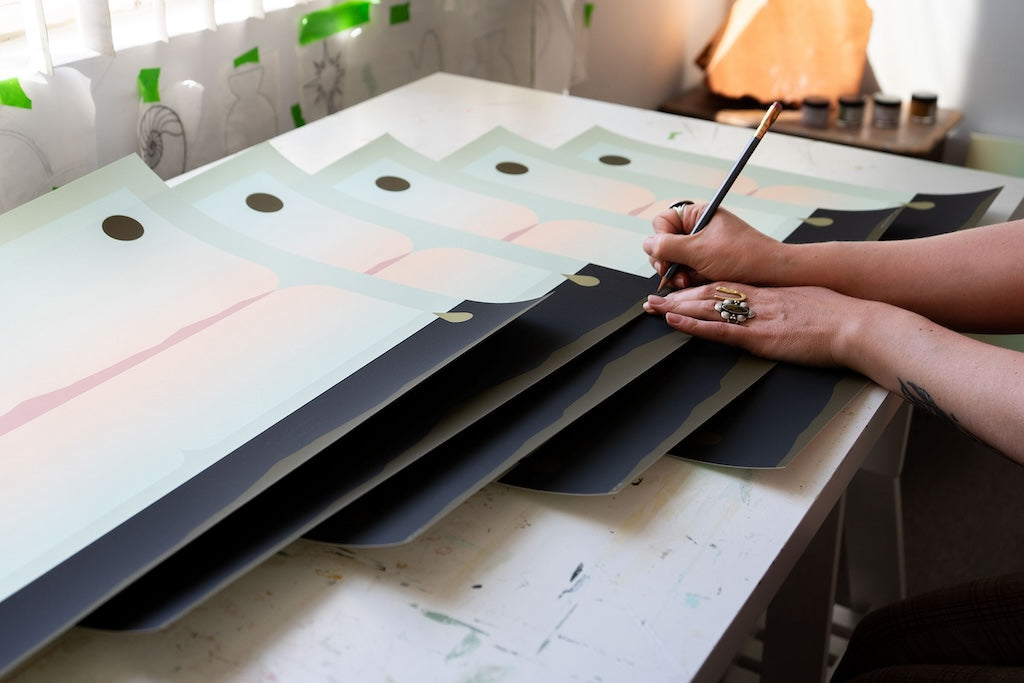 Madeleine Tonzi signing prints of a landscape in neutral greens, blues, and pinks with a black border at the bottom