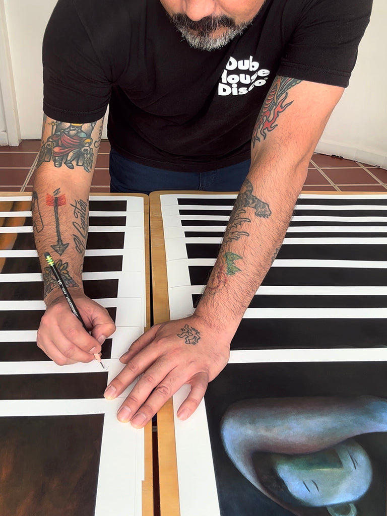 Carlos Rodriguez (man with arm tattoos in black tshirt) signing prints of four men laying on the floor and cuddling one another. The background is in dark blue.