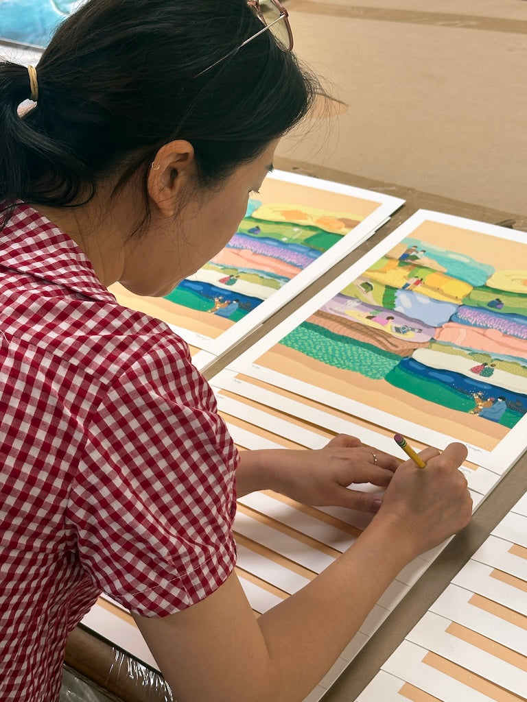 Woman in a checkered shirt signing a series of colorful prints.
