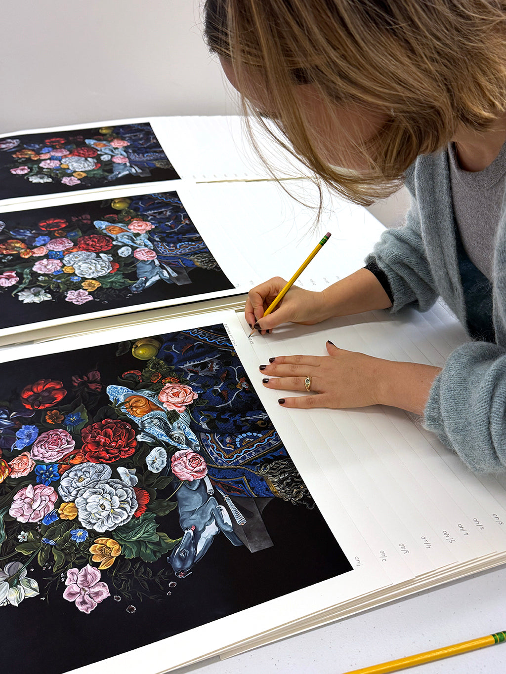 Sabrina Bockler signing print of a still life featuring a bouquet of flowers, a grey dog, and an ornately patterned blue cloth against a black background by Sabrina Bockler