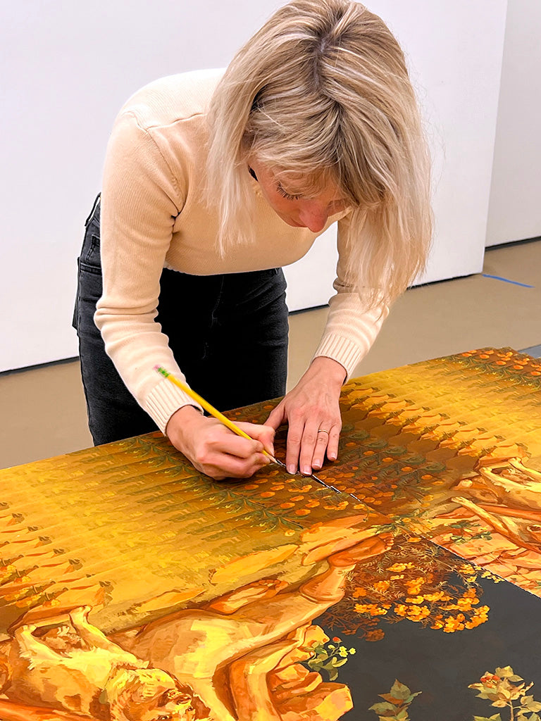 Rachel Gregor (blonde white woman) signing prints in yellow and green tones of nude woman sitting on ground with back to viewers. Two dogs near her, one snarling while the other is looking at the viewer