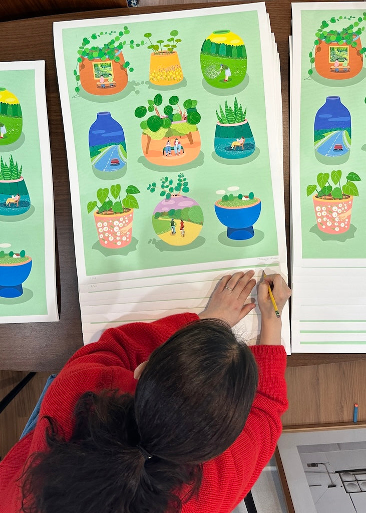 Danym Kwon signing (shot from above of woman with dark hair in red sweater) signin prints of vases with plants on green background. Each vase has details such as people in fields, cars driving down lanes, people on bicyclyes