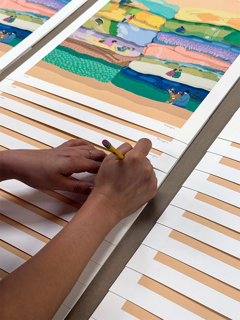 Person signing a landscapes with figures set within laundry stacks with a pencil 