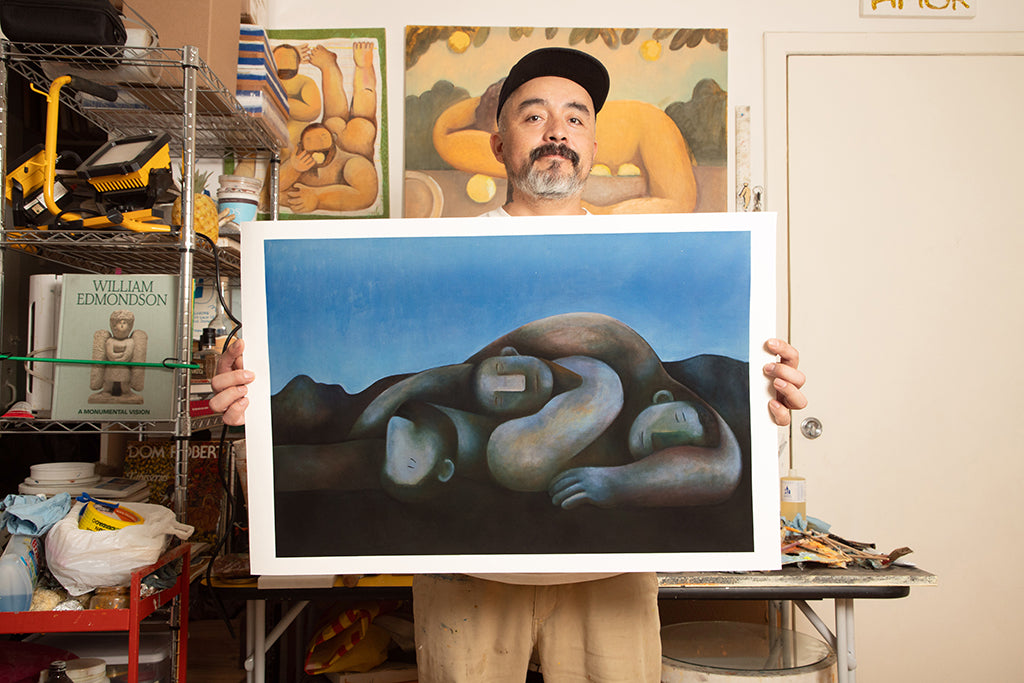 Carlos Rodriguez (man in black baseball hat) holding up print of four men laying on the floor and cuddling one another. The background is in dark blue.