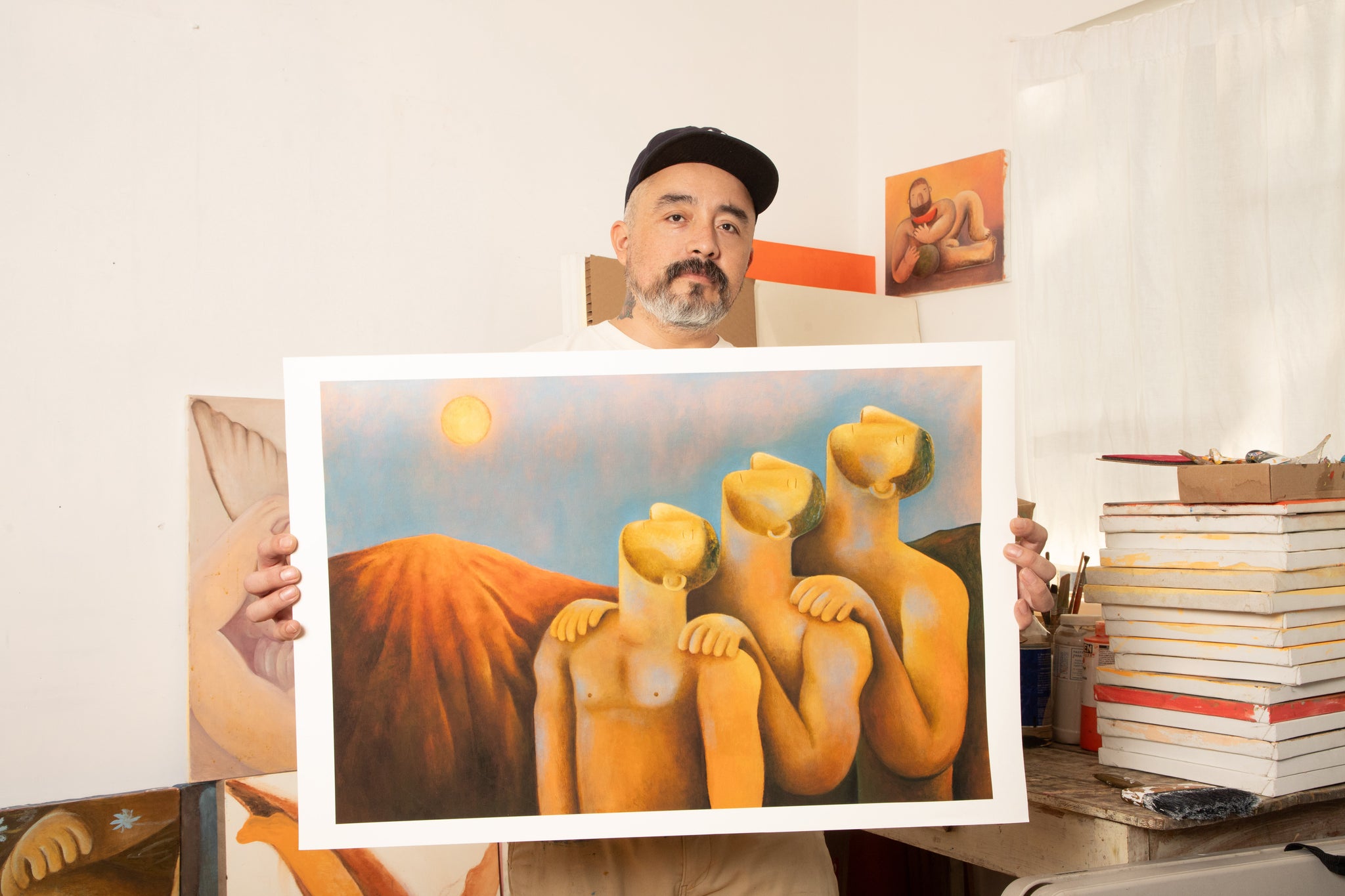 Carlos Rodriguez (man in black baseball hat) holding up print of three men in nudity and have their head tilted upwards. They stood in line with their hands on another's shoulder in front of the mountain and sun on the left side