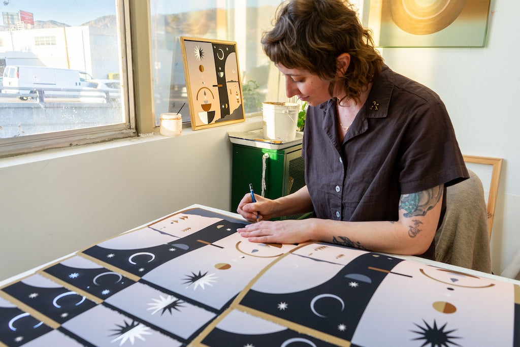 white woman in brown short sleeve shirt sitting by window signing winter solstice inspired print in blacks, earthy and pink tones