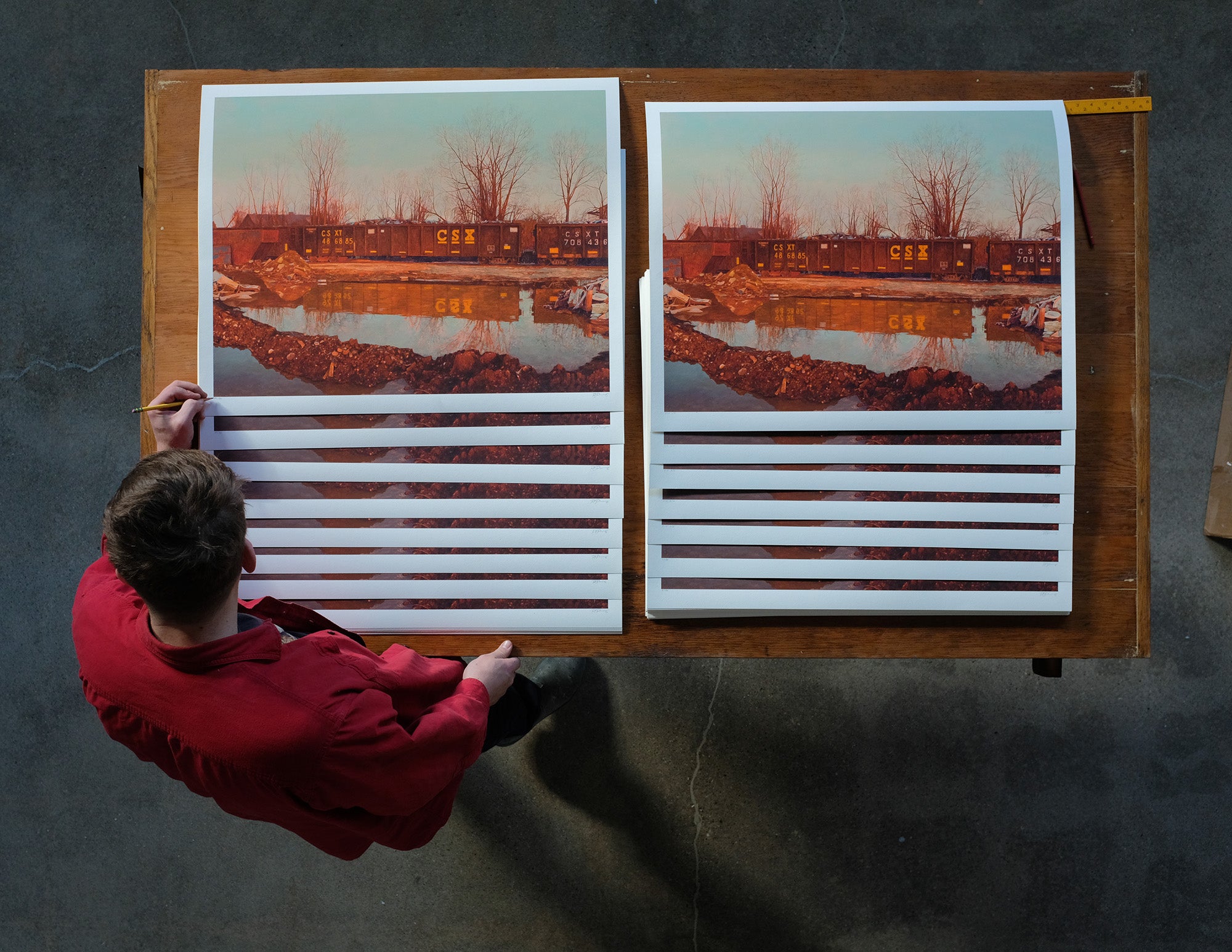 A person in a red shirt stands at a wooden table, inspecting two limited edition Pat Perry Ameliorator prints, depicting a train by trees and water, with ruled strips of paper placed below each print.