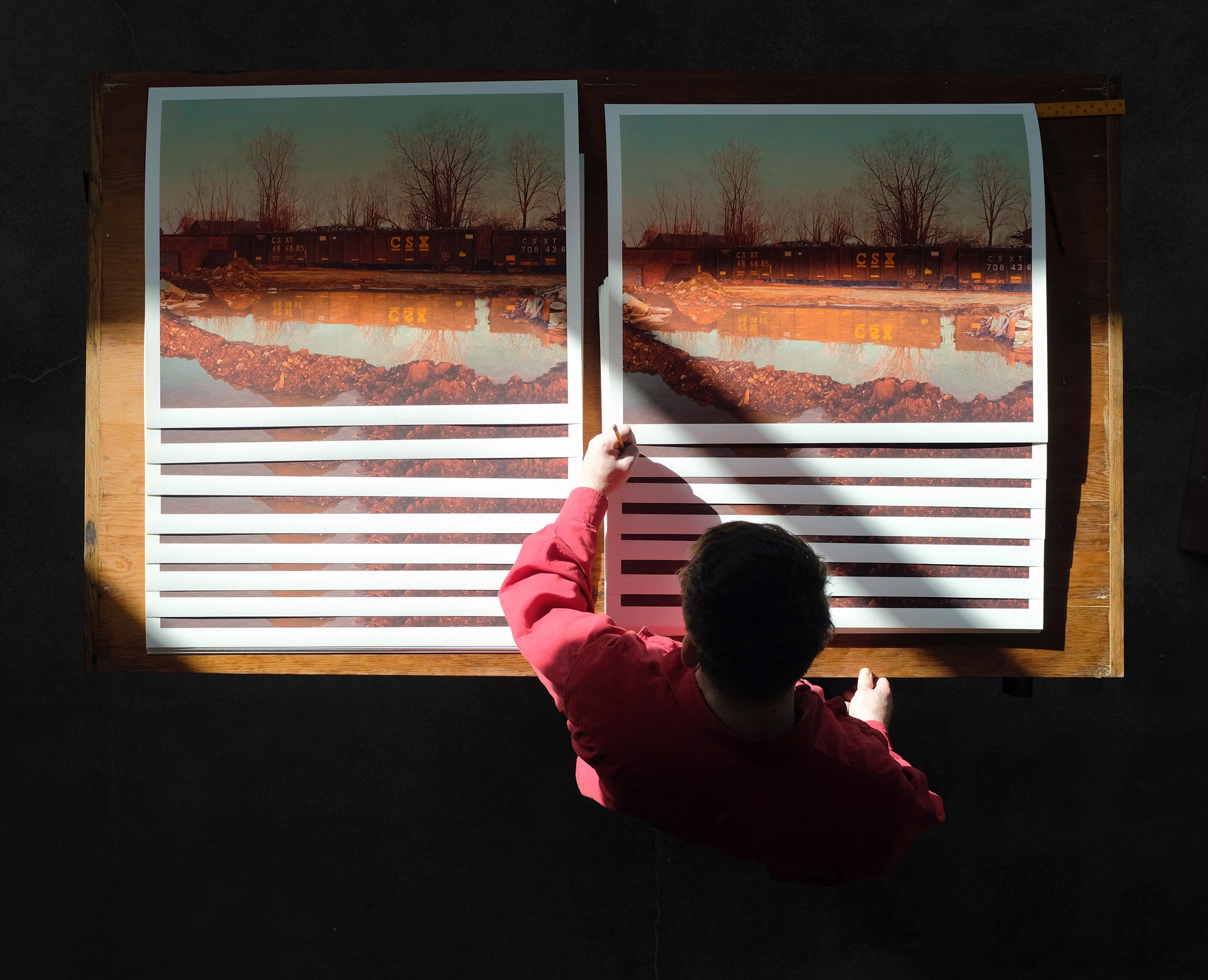 A person in a red shirt examines two identical Pat Perry Ameliorator archival pigment prints side by side on a wooden table, with light shining on the workspace from the left.