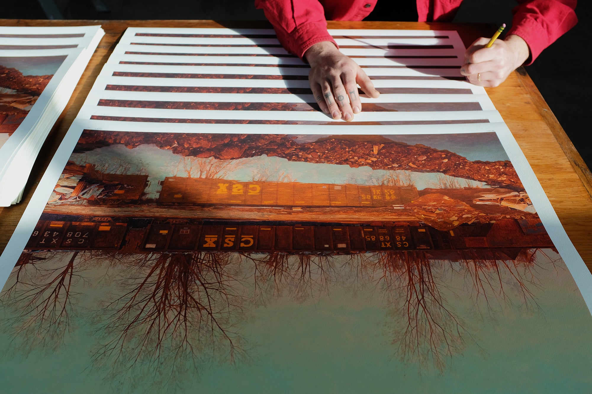 A person in a red shirt signs Pat Perrys limited edition Ameliorator archival pigment prints, which feature train cars amid leafless trees and rocky terrain. The prints appear upside down on the wooden table. Brand: Pat Perry.