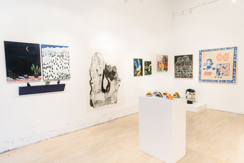A gallery room with artworks on white walls features paintings of landscapes, abstract art, and a large tree stump piece. Liz Hernandezs striking Floral Times, an acrylic on canvas from 2017, measures 28 x 26 inches. A white pedestal displays colorful sculptures on the light wood floor.